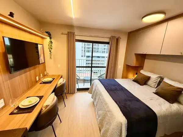 O mesmo studio pronto: cama de casal arrumada, painel de madeira, TV e mesa para dois